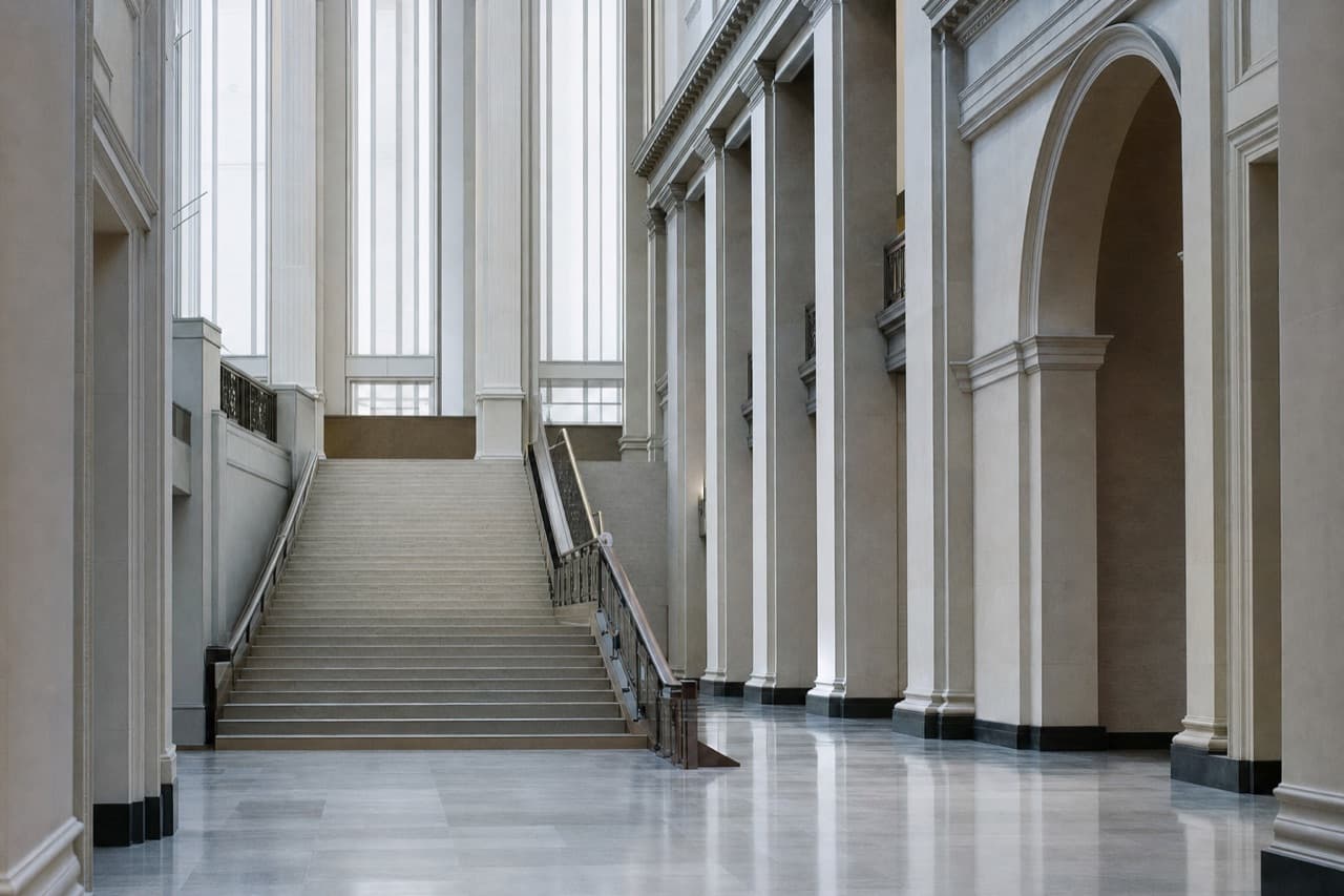A wide stone staircase rising beneath tall windows inside a government building.