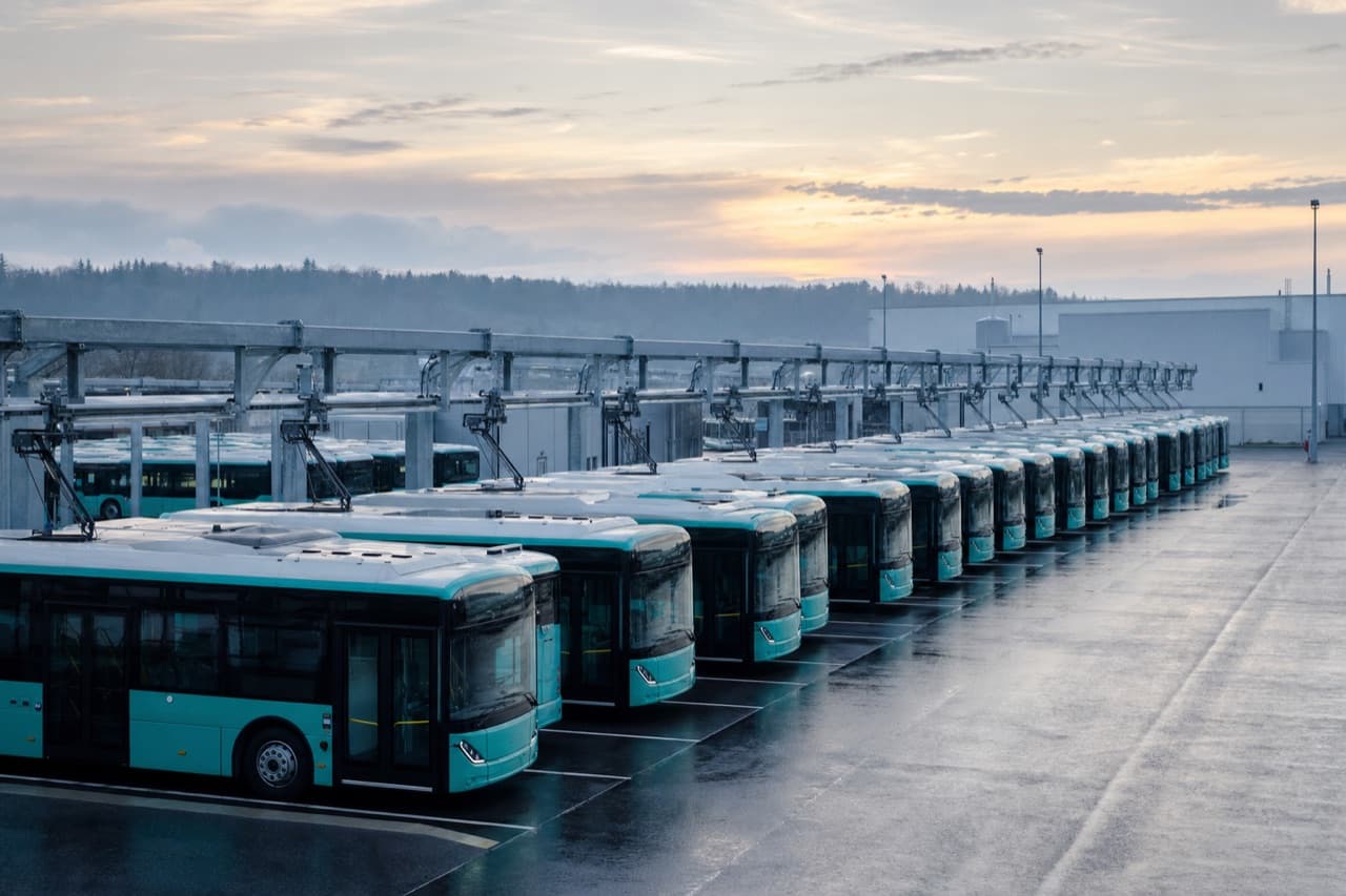 Rows of electric city buses parked under overhead charging gantries at dawn.